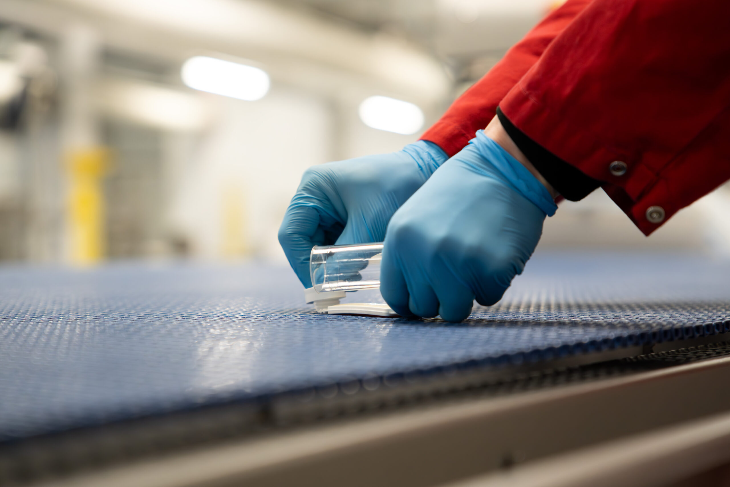 Hender med blå engangshansker tar en miljøprøve med svaber på et transportbånd i et næringsmiddelanlegg.
