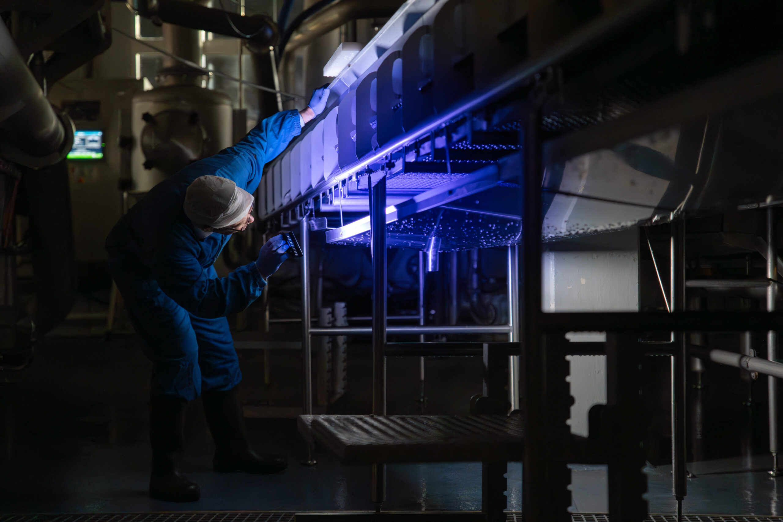 Renholdsarbeider i blått verneutstyr inspiserer produksjonsutstyr i et næringsmiddelanlegg, belyst av blått lys i et industrielt miljø.